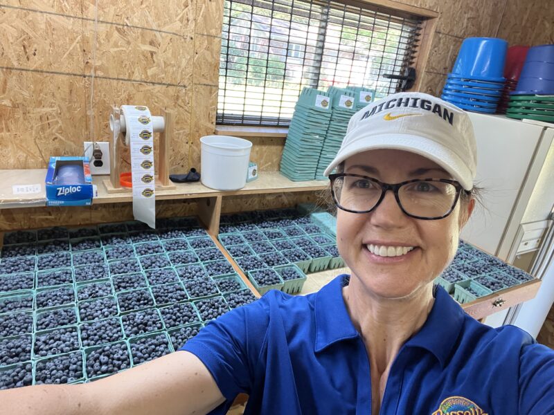 Woman smiling in a blueberry packing facility surrounded by containers filled with fresh blueberries, with tools and supplies for packaging in the background.