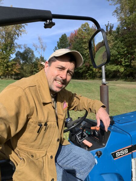 Man smiling while sitting on a blue tractor, wearing a tan jacket with an American flag patch and a cap, surrounded by autumn foliage. The tractor's dashboard is visible, showcasing controls and instruments.