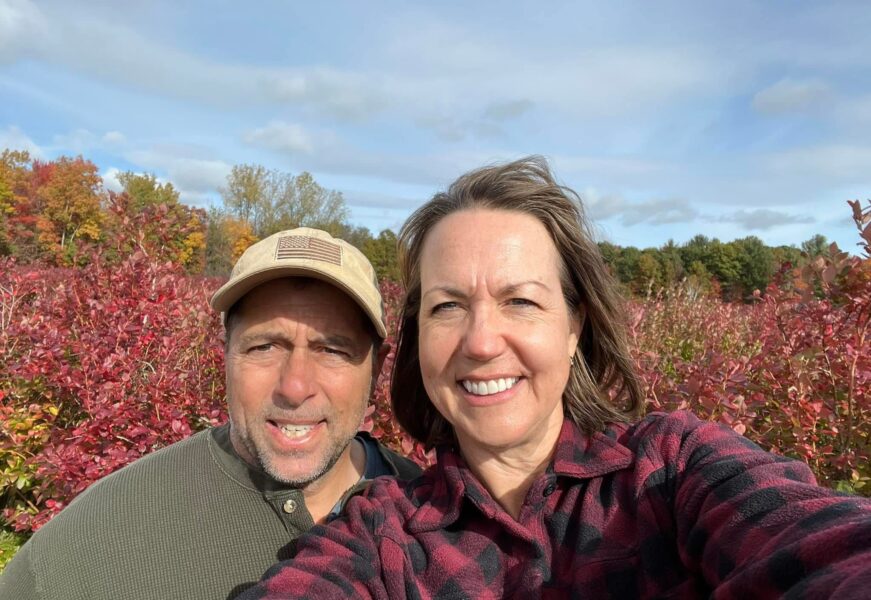 A couple enjoying a sunny day at a blueberry farm, surrounded by vibrant fall foliage and berry bushes in shades of red and green.