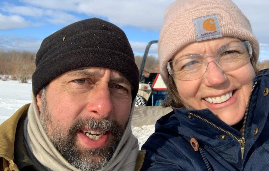 Two people smiling for a selfie in a snowy outdoor setting, wearing winter clothing and protective eyewear. The background features a blue sky with clouds and hints of trees.