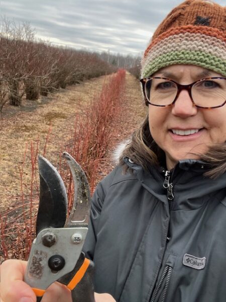 Woman holding pruning shears in a berry field during winter, wearing a warm hat and jacket, with rows of bare bushes in the background.
