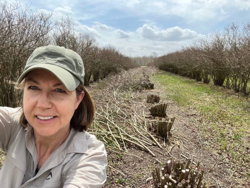 Woman in a cap smiling for a selfie in a field lined with pruned bushes and cut branches, under a partly cloudy sky.