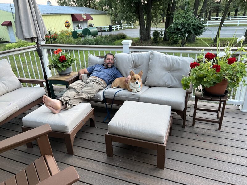 Man relaxing on a patio couch with a corgi dog, surrounded by potted flowers and outdoor furniture, in a scenic garden setting.